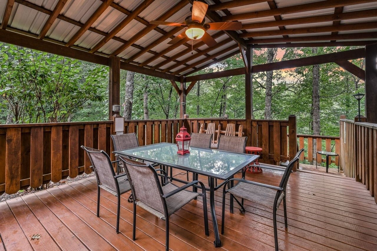 An outdoor covered patio with a glass table and chairs, surrounded by wooden railings and a view of trees.