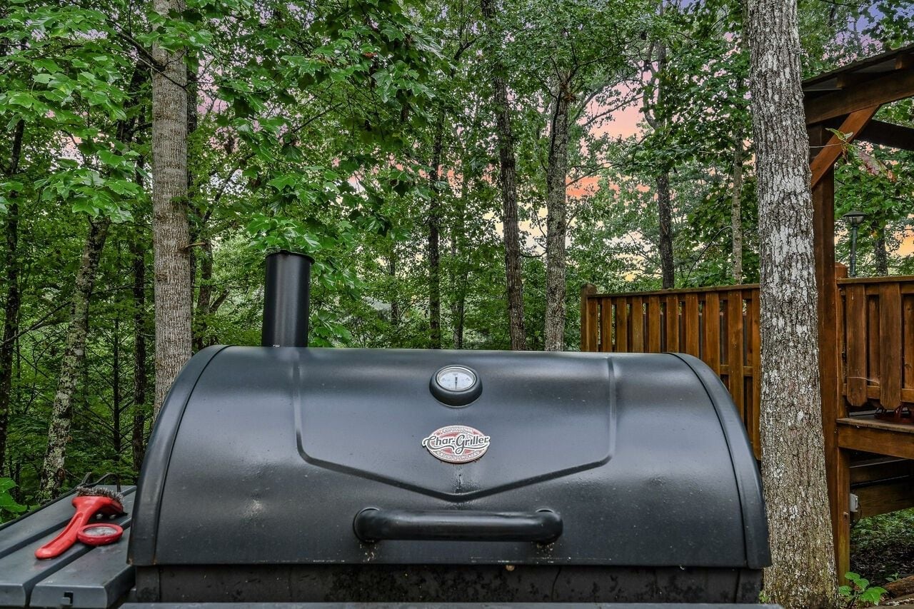 A charcoal grill with a temperature gauge, positioned outdoors with trees in the background.
