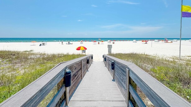 A wooden boardwalk leading to a beach with umbrellas and a flag. - Windward Pointe 502-Beachfront 3bd/2ba-Sleeps 8-Heart of Orange Beach (Orange Beach)