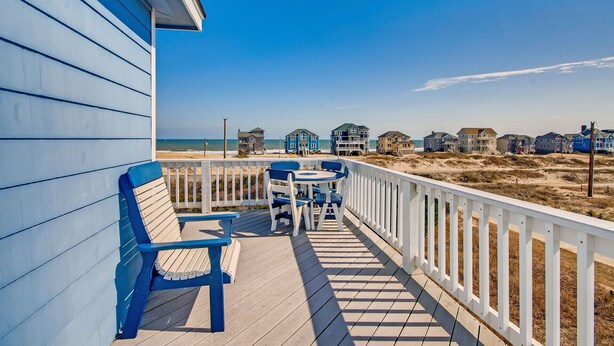 A wooden deck with blue chairs and a table, overlooking a beach with several houses. - Premier Semi-Oceanfront w/ Pool, Hot Tub, Elevator, Theater, Bocce/Putting Court (Rodanthe)