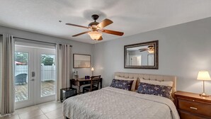 A bedroom with a bed, a desk, a chair, a ceiling fan, a mirror, and a sliding glass door leading to an outdoor area.
