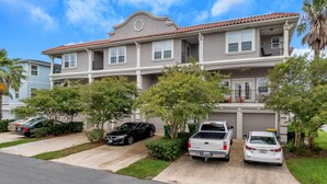 A two-story residential building with a red-tiled roof, multiple windows, and a balcony on the upper floor. There are three vehicles parked in the driveway, including a white pickup truck and a black sedan.