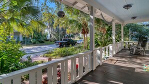 A covered porch with a white railing, surrounded by lush greenery and a view of a street with parked cars. - Sunny Side Cottage walkable to downtown w/ garden (St Augustine)