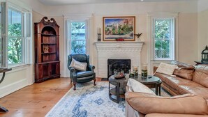 A living room with a fireplace, leather sofa, wooden bookshelf, and a chandelier. - Sunny Side Cottage walkable to downtown w/ garden (St Augustine)