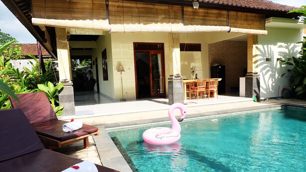Una piscina con un flotador de flamenco rosa, sillas de madera para tomar el sol y un área exterior techada con una mesa y sillas.