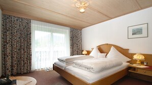 Quarto de hotel com cama de madeira, duas mesas de cabeceira, luminária, quadro emoldurado na parede e janela com cortinas.