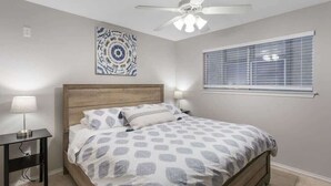 A bedroom with a wooden bed, a nightstand, a ceiling fan, a window with blinds, and a wall art piece. - Relax by the River | Shared Pool | Labor Day Ready (New Braunfels)