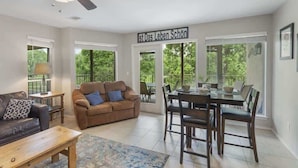 A spacious living room with a ceiling fan, a brown sofa, a dining table with chairs, and large windows offering a view of greenery. - Relax by the River | Shared Pool | Labor Day Ready (New Braunfels)