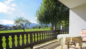 A balcony with a view of a green field and mountains, a table with a striped tablecloth, and wicker chairs. - Apartment 2, 55sqm, terrace, kitchen extra, 1 bedroom, max 2 pers., WLAN (Ruhpolding)