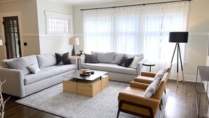 Una sala de estar moderna con un sofá gris, una mesa de centro de madera, una mesita auxiliar con una lámpara, una lámpara de pie y un ventanal con cortinas translúcidas.