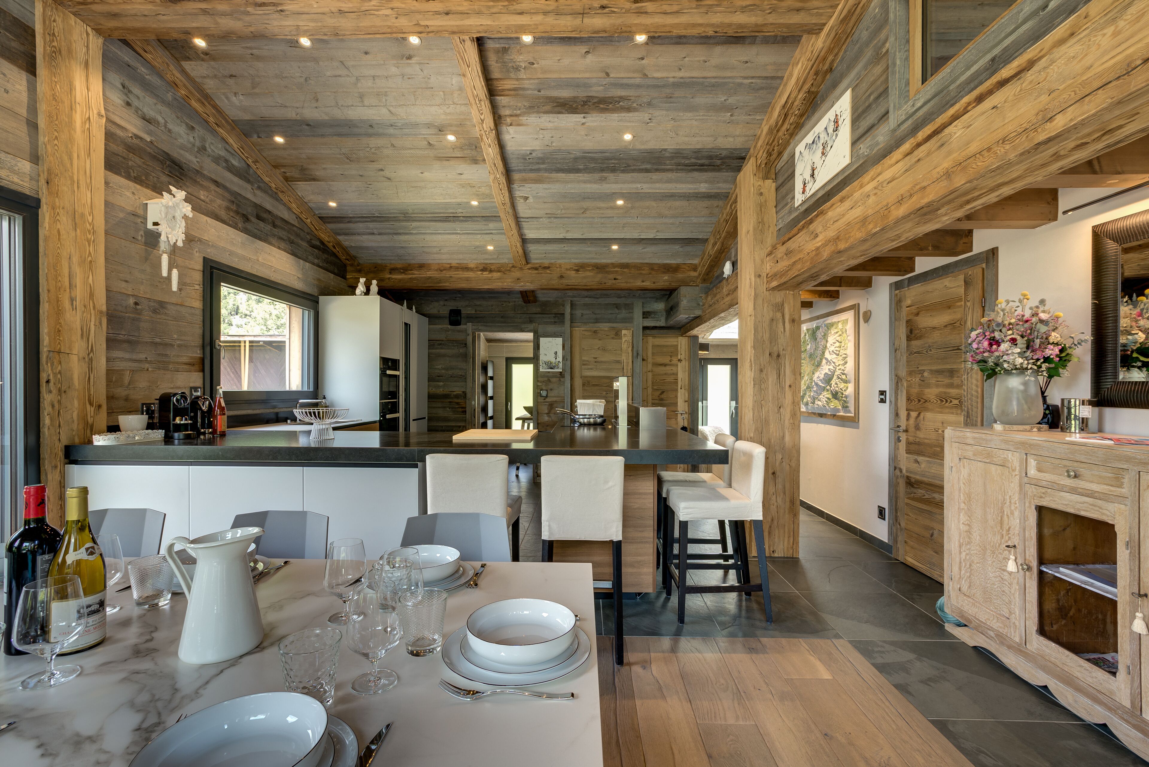 Una cocina moderna con vigas de madera, una isla central y un comedor con sillas blancas y una mesa de mármol.