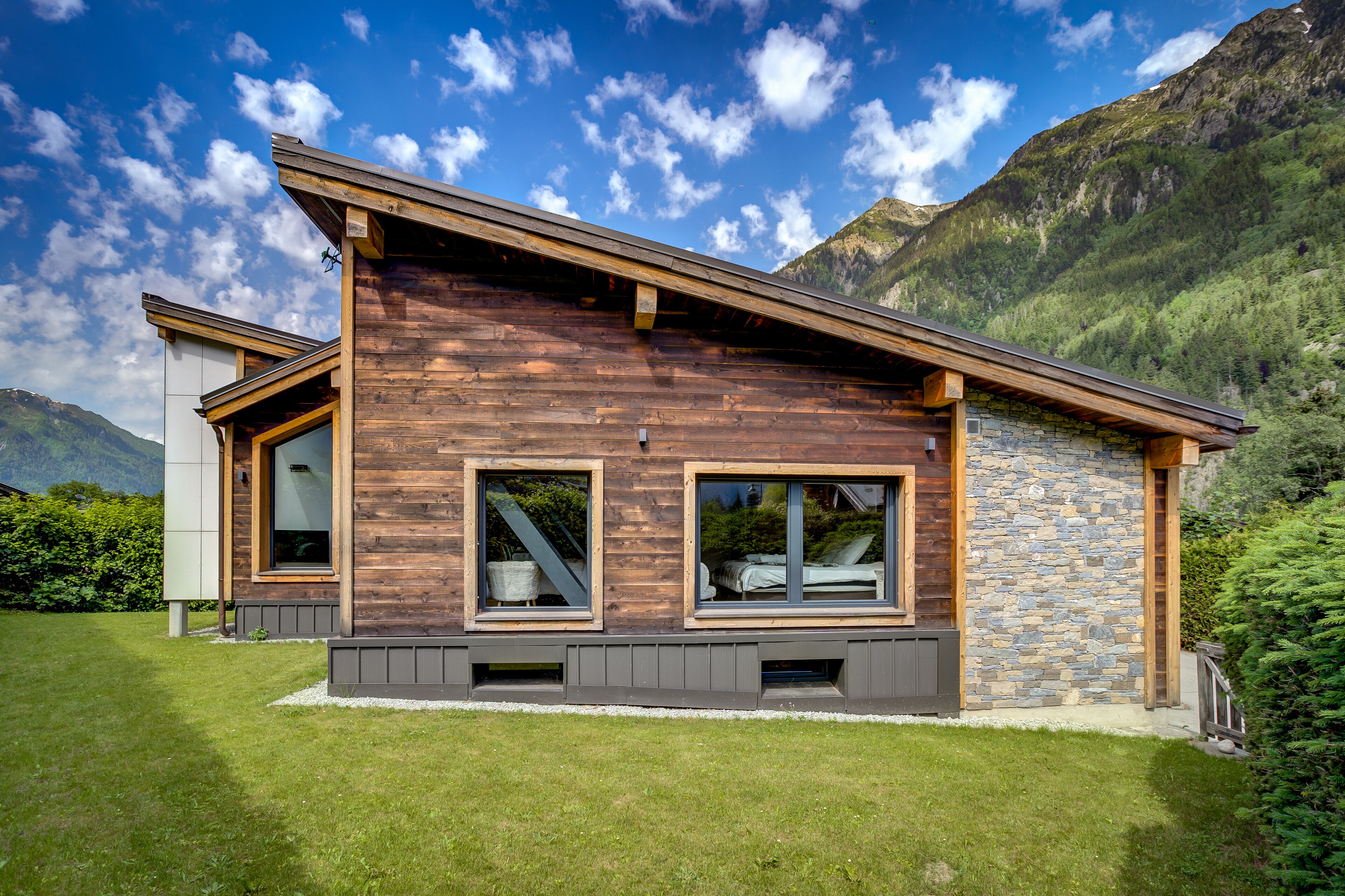 A modern house with a sloped roof, large windows, and stone walls, surrounded by a well-maintained lawn and mountains in the background.