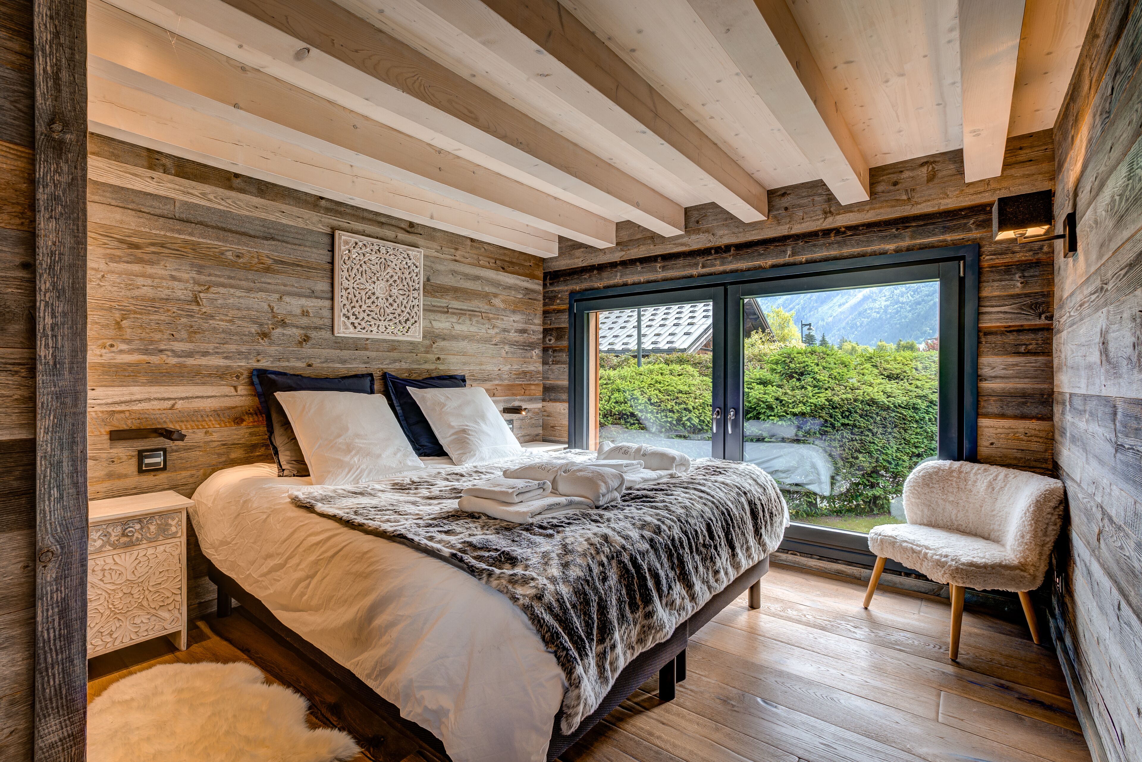 Un dormitorio con una cama grande, paredes de madera, una ventana con vistas a zonas verdes y una silla junto a la ventana.