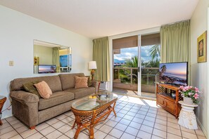 A living room with a sofa, a glass coffee table, a TV on a wooden stand, and a balcony with a view of palm trees.