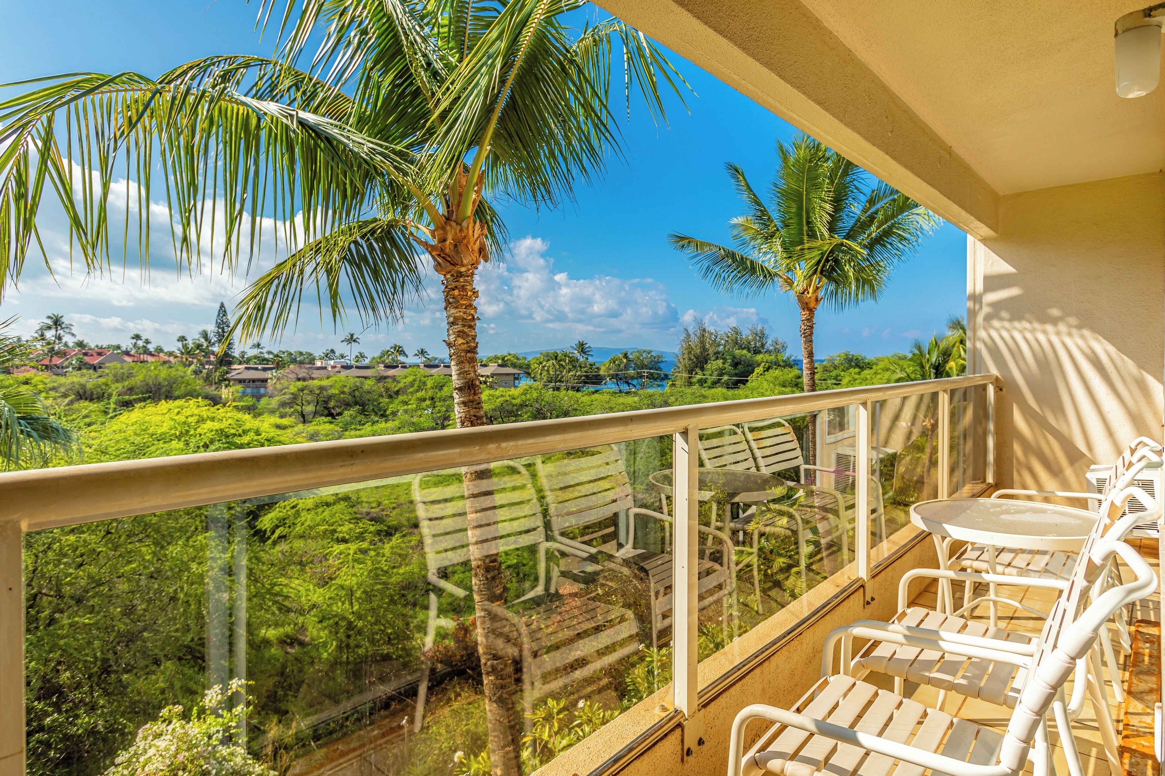 Un balcón con sillas y una mesa blancos, con vistas a un paisaje verde exuberante con palmeras y casas.