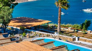 A pool area with wooden loungers, a palm tree, and a view of the sea and beach. - Karkos Beachfront Villa  Private pool (Mykonos)
