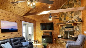 A cozy living room with a black leather sofa, a grey armchair, a wooden coffee table, and a stone fireplace. - Cub Cabin on Massanutten Resort- Fully Renovated Log Cabin- Hot Tub & Fire Pit (Massanutten)