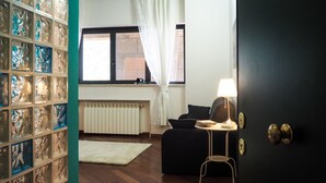 A room with a wooden floor, a white radiator, a black sofa, a small table with a lamp, and a glass display case with various items. - Amba Alagi Apartment (citra 010025-LT-1799) (Genova)