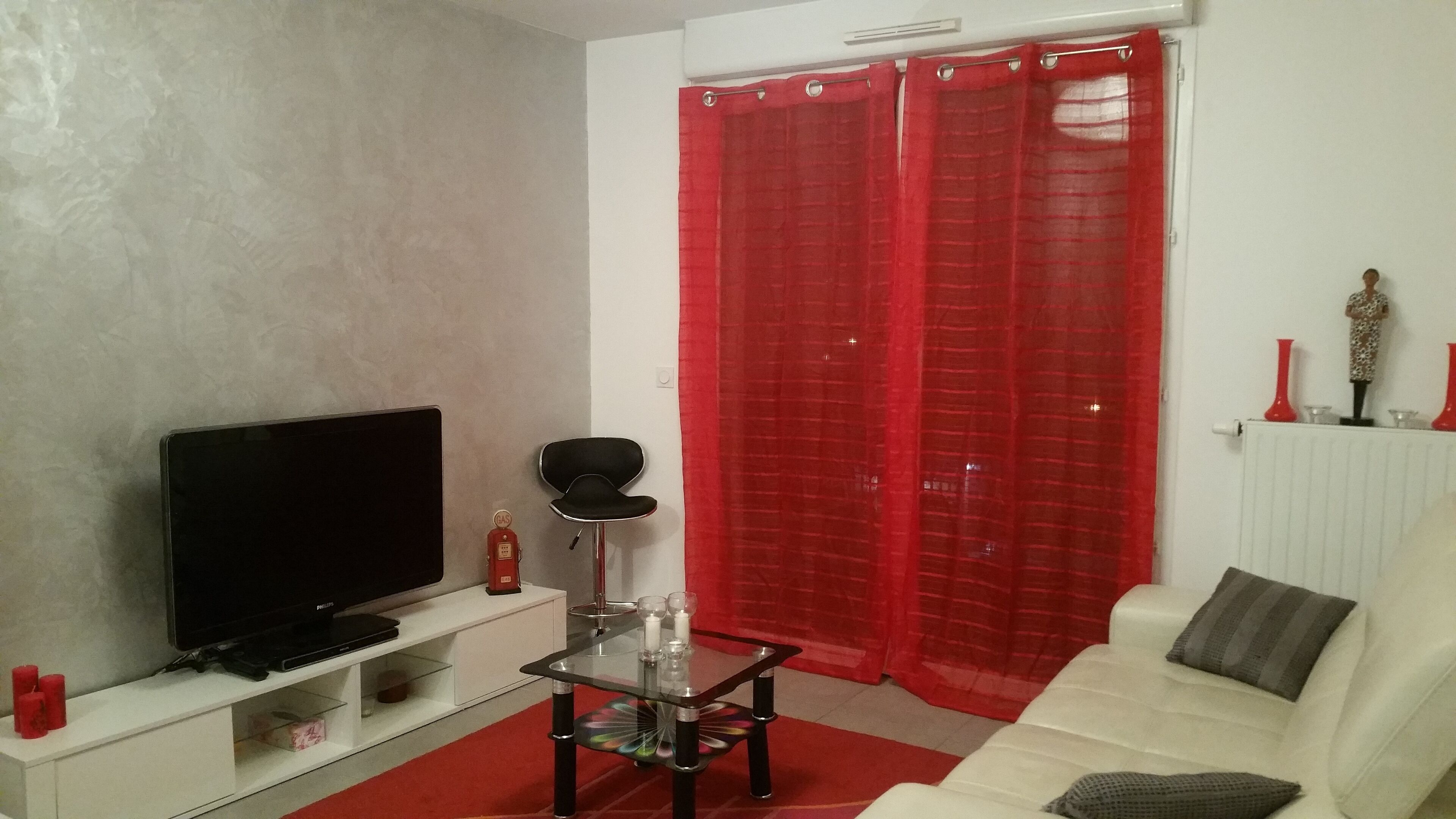 A living room with a flat-screen TV on a white stand, a glass coffee table with a colorful base, a red curtain, and a white sofa with a gray cushion.