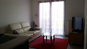 A living room with a beige sofa, a glass coffee table, a red rug, a TV on a stand, and a window with sheer curtains.