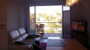 A modern living room with a sofa, a dining table, and a TV displaying a cartoon. A balcony with a view of trees and houses is visible through a glass door.