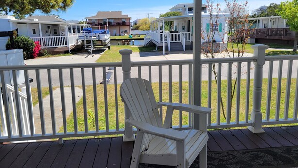Une véranda couverte avec un fauteuil Adirondack blanc, une vue sur une rue résidentielle bordée de maisons et d’un petit plan d’eau, et un ciel bleu dégagé.