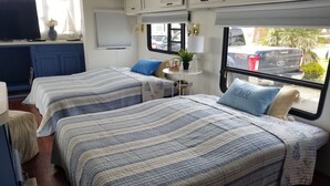 Une chambre confortable dans un camping-car, avec deux lits, une petite table agrémentée d’une plante et une fenêtre donnant sur les voitures garées.