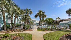 A pool area with palm trees, a concrete walkway, and a building with a green roof. - Gulf view condo - Now Incl. Beach Service  (Miramar Beach)