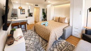 A modern hotel room with a bed, desk, kitchenette, and a ceiling fan.