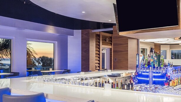 A modern bar area with a long counter, bar stools, and a variety of bottles and glasses. - Beach Vacay With View! Direct Beach Access, Pool, Bar, Restaurant! (Fort Myers Beach)
