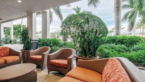 A covered outdoor seating area with wicker furniture and orange cushions, surrounded by lush greenery and palm trees. - Beach Vacay With View! Direct Beach Access, Pool, Bar, Restaurant! (Fort Myers Beach)