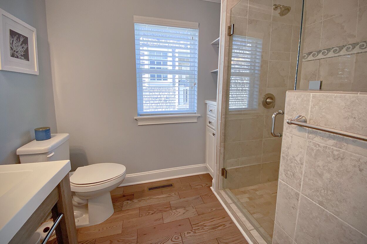 A bathroom with a toilet, a window with blinds, a shower with a glass door, and a sink with a countertop.