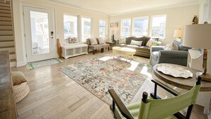 A spacious living room with hardwood floors, a large rug, and multiple seating areas including a sofa, chairs, and a bench. - Beautifully updated cottage with ocean views, only steps to the beach! (Wrightsville Beach)