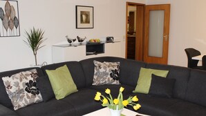 Ein modernes Wohnzimmer mit einem dunkelgrauen Ecksofa, grün gepolsterten Kissen, einem weißen Couchtisch mit einer Vase voller gelber Blumen und einem gerahmten Bild an der Wand.