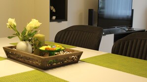 Ein Essbereich mit einem für zwei Personen gedeckten Tisch, der ein dekoratives Tablett mit Blumen und einer Kerze zeigt. Im Hintergrund ist ein Fernseher an der Wand befestigt.