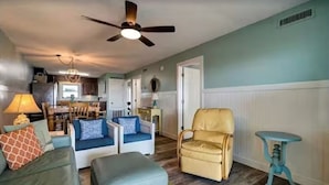 Un salon avec un ventilateur de plafond, un coin repas avec une table et des chaises, et une cuisine à l’arrière-plan.