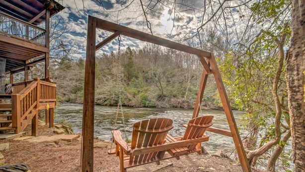 Una silla mecedora de madera junto a un río, una terraza de madera y una cabaña al fondo.