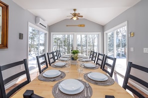 Enjoy meals in this spacious dining room with a long table, ceiling fan, and air conditioning unit, offering a view of the surrounding snowy landscape.