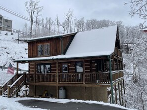 A wooden cabin with a balcony and a covered porch, surrounded by snow-covered trees.