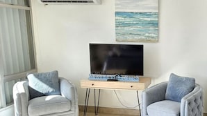Una sala de estar con dos sillones grises, un televisor de pantalla plana sobre un pedestal de madera, y un cuadro del océano en la pared.