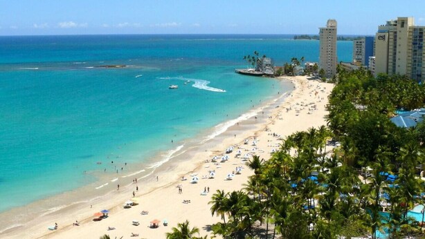 Una playa con aguas turquesas cristalinas, una orilla de arena y varias palmeras. Se observan edificios al fondo, probablemente hoteles o complejos turísticos.