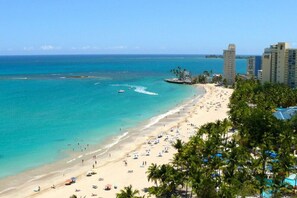 A beach with clear turquoise waters, a sandy shore, and several palm trees. There are buildings in the background, likely hotels or resorts.
