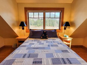 Un dormitorio con una cama grande, dos mesitas de noche, un ventanal con vistas a árboles y dos lámparas de pared.
