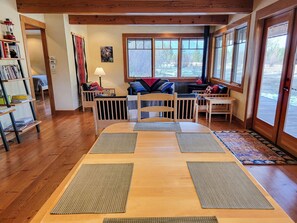 Una habitación acogedora con suelo de madera, una mesa de comedor con tapetes, una estantería, un ventanal con vistas y un sofá con cojines.