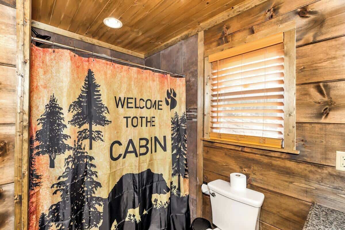 A cabin interior with a wooden ceiling, a toilet, a window with blinds, and a curtain with a forest and animal print.