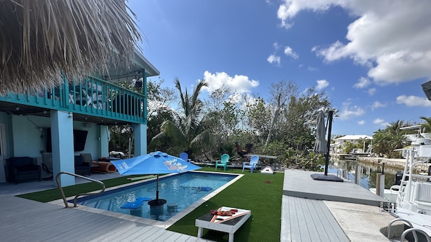 A pool area with a blue umbrella, a deck, and a thatched roof structure. - The most setup home in BPK TIKI HUT,POOL ,GRILL ,CANAL FRONT, FREE UNLIMITED ICE (Gordon road)