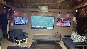 A cozy outdoor seating area with multiple screens displaying sports events, comfortable seating, and a rustic thatched roof. - The most setup home in BPK TIKI HUT,POOL ,GRILL ,CANAL FRONT, FREE UNLIMITED ICE (Gordon road)