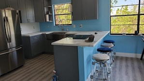 A modern kitchen with grey cabinets, a stainless steel refrigerator, a sink, and a bar area with stools. - The most setup home in BPK TIKI HUT,POOL ,GRILL ,CANAL FRONT, FREE UNLIMITED ICE (Gordon road)