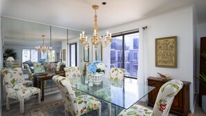 Een eetruimte met een glazen tafel, stoelen bekleed met bloemenprint, een kroonluchter en uitzicht door grote ramen.