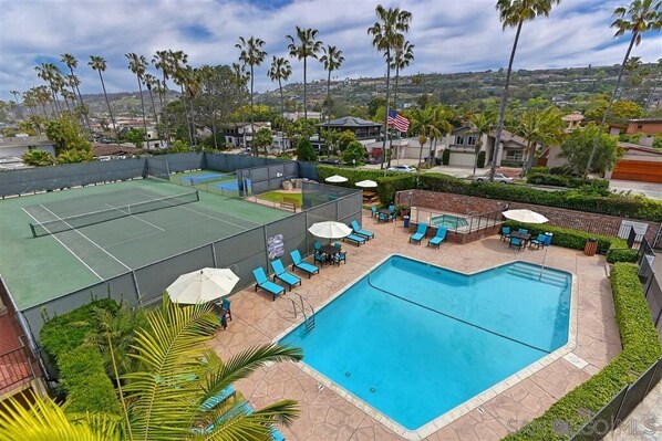 A rooftop pool area with a tennis court, lounge chairs, and a swimming pool.